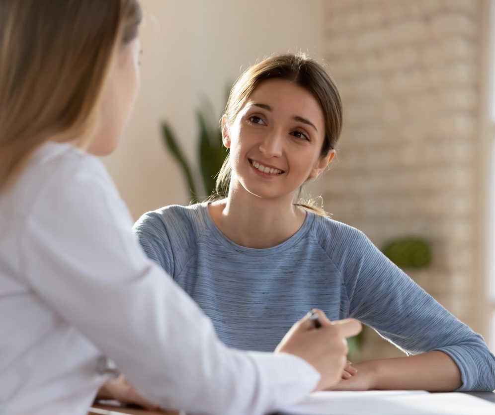 Smiling millennial woman visit female doctor consulting about health complains, happy satisfied young client talk discuss treatment prescription with GP or physician at appointment in hospital